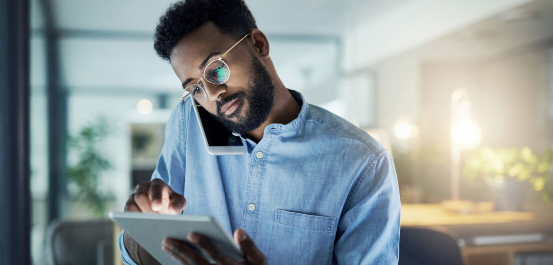 Ein junger Mann im Büro bedient ein Tablet in der Hand, während er ein Smartphone am Ohr eingeklemmt hat