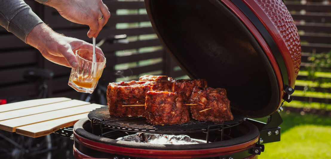 Roter Keramikgrill mit Rippchen auf dem Rost, die gerade von einem Mann mit Marinade bestrichen werden