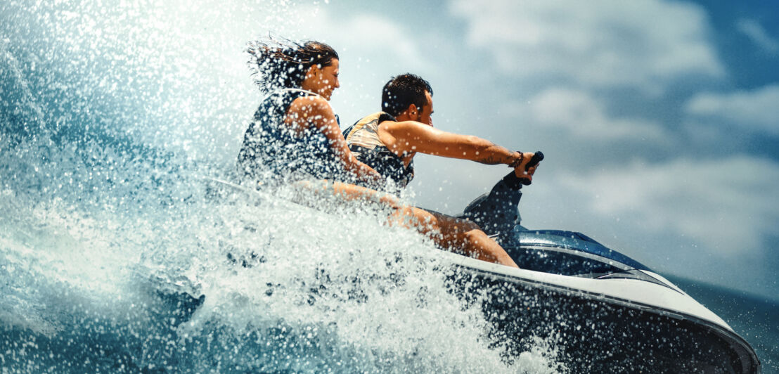 Zwei Personen fahren auf einem Jetski, vor ihnen die Gischt