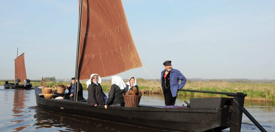 Kleines Holzschiff mit Stoffsegel und fünf Passagieren schippert über einen kleinen Fluss