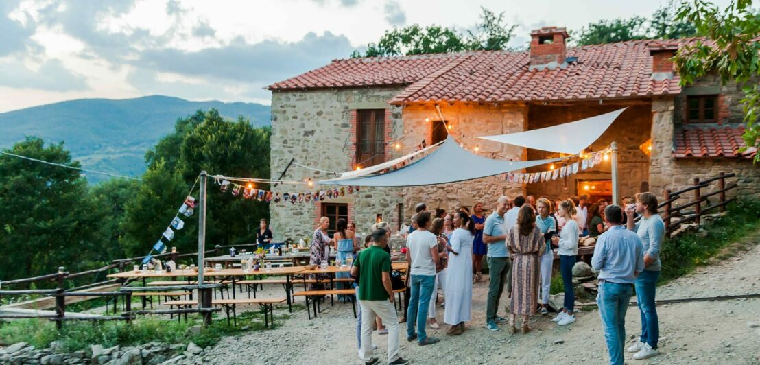 Mehrere Personen stehen um Biertische vor einem Steinhaus, im Hintergrund Berge