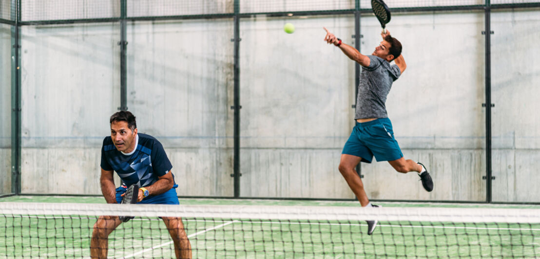 Zwei Männer spielen Padel-Tennis, von denen einer gerade einen Aufschlag macht.