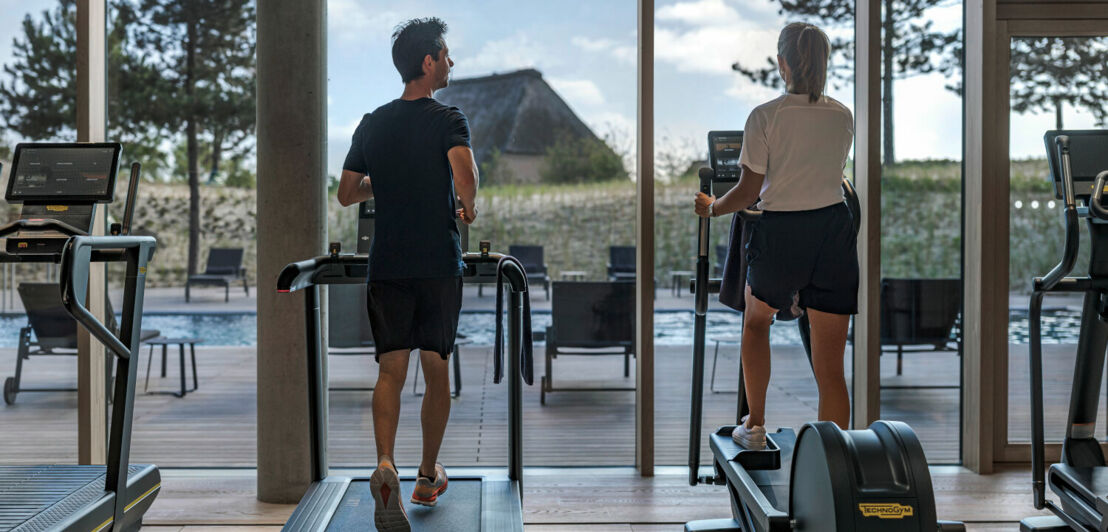 Rückansicht zweier Personen, die auf Fitnessgeräten mit Blick in die Dünen trainieren