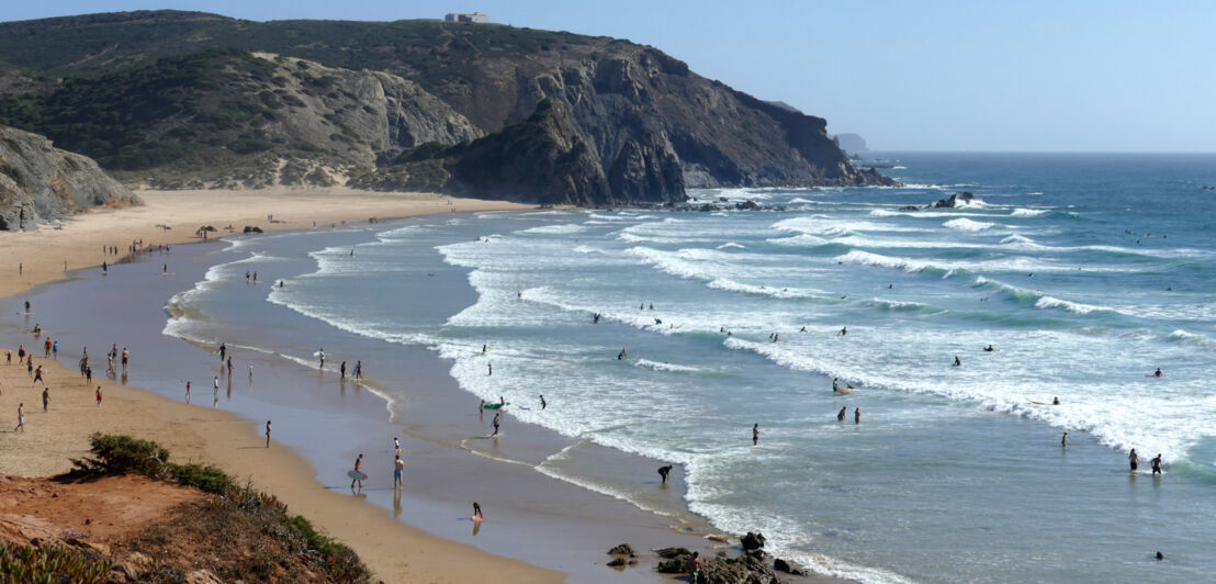 Panorama einer Bucht mit Sandstrand und Personen im Wasser, im Hintergrund Felsen