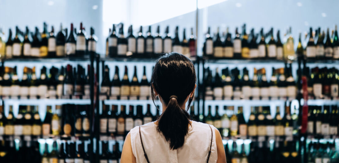 Eine dunkelhaarige Frau von hinten, die vor einem Weinregal steht.