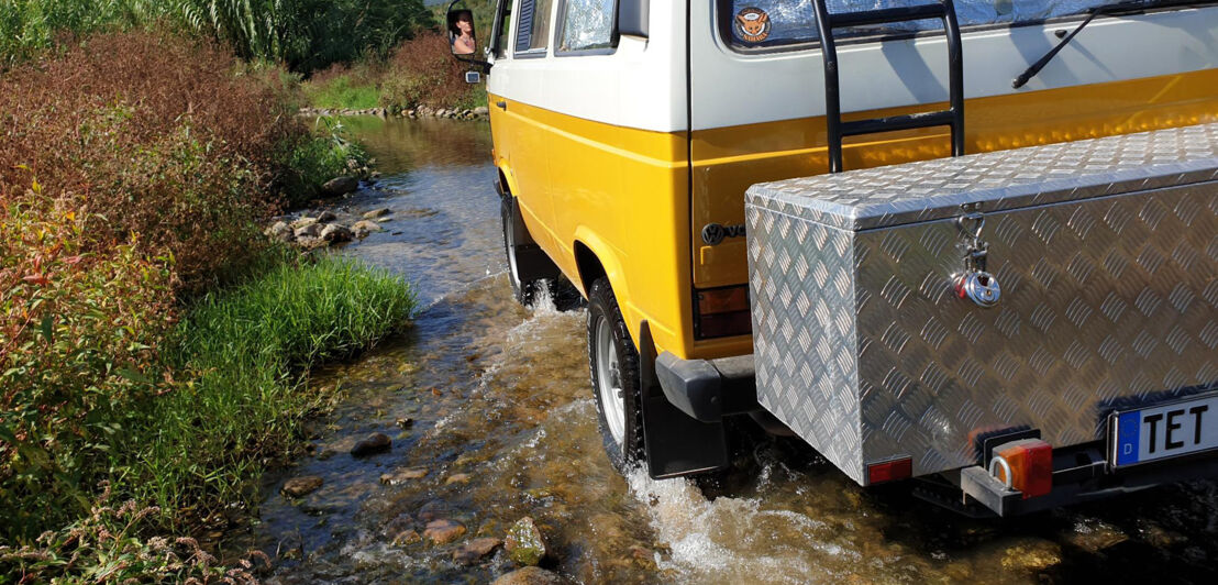 Heckansicht eines gelben VW Bus mit großer Heckbox, der durch ein Flussbett fährt