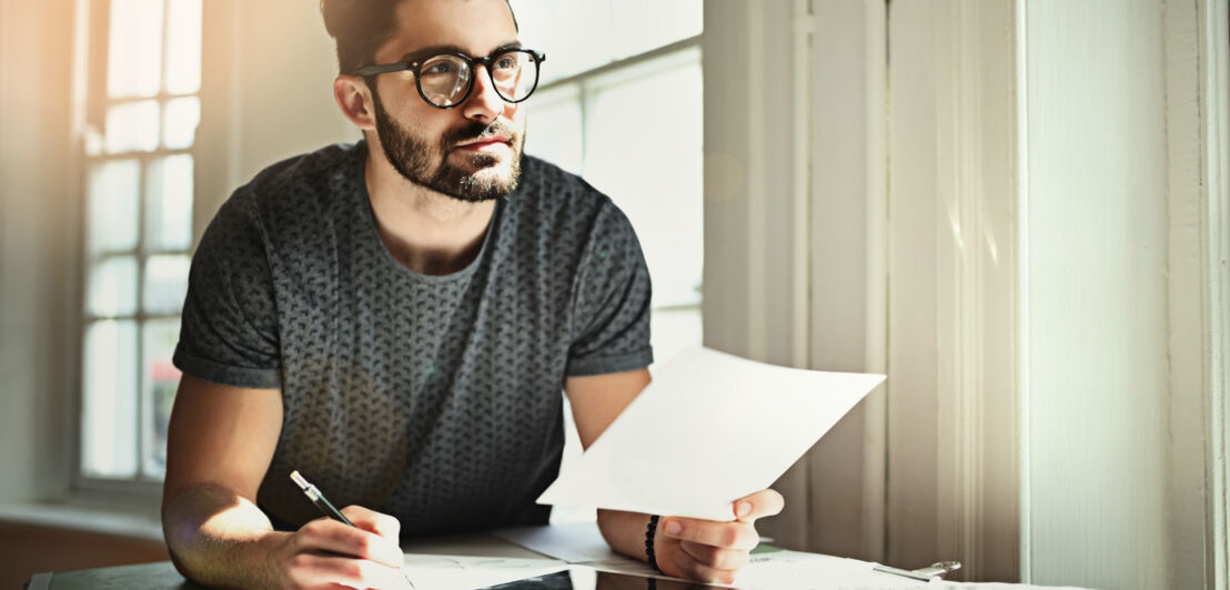 Eine Person lehnt auf einem Schreibtisch und hält dabei einen Stift sowie ein Blatt Papier in seinen Händen