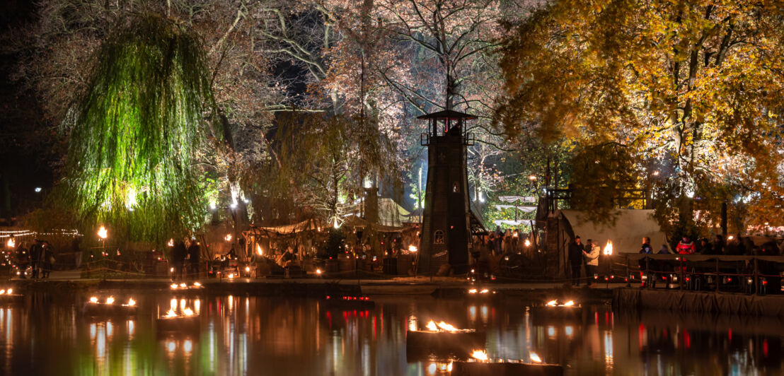 Ein See bei Nacht mit schwimmenden Fackeln und einem beleuchteten Ufer, das von Zelten gesäumt wird