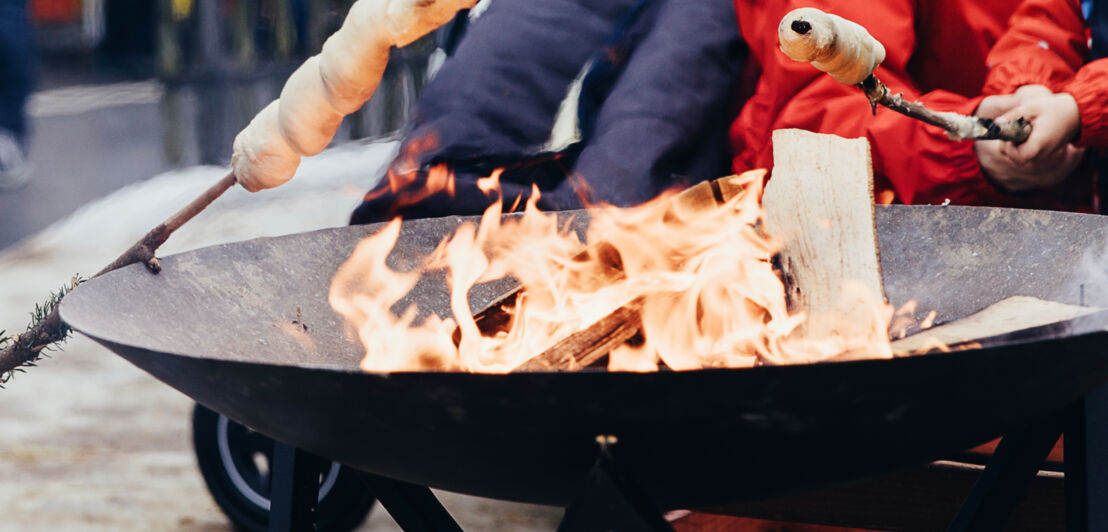 Feuerschale mit einem lodernden Feuer, über dem Kinder Stockbrot backen