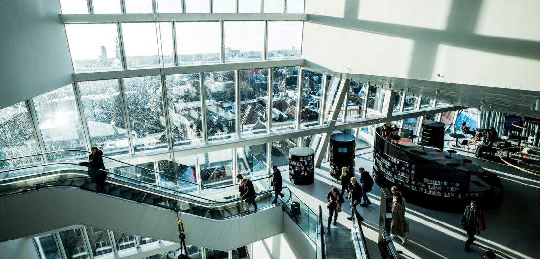 Innenaufnahme eines modernen Gebäudekomplexes mit Personen auf Rolltreppen und Panoramablick über eine Stadt