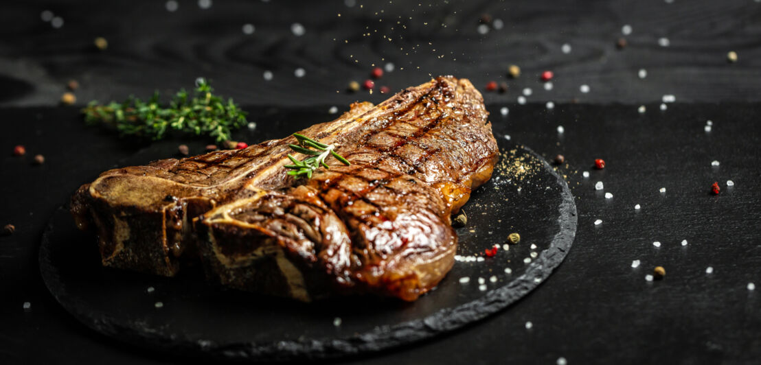 Ein T-Bone gegrilltes Rindfleischsteak auf einem schwarzen Teller, garniert mit bunten Pfefferkörnern