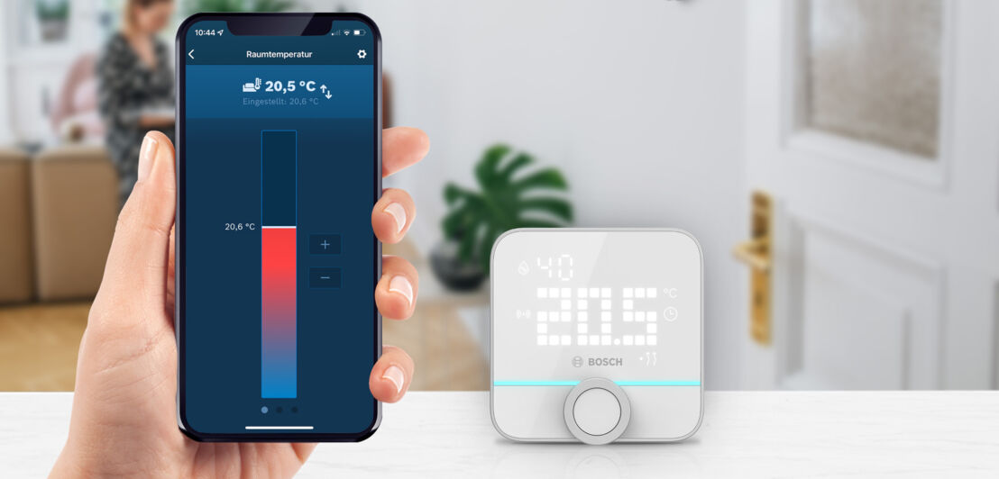 Eine Hand hält ein Smartphone mit einer Temperaturanzeige, daneben ist ein Raumthermostat in weiß zu sehen