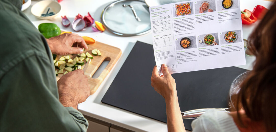 Zwei Personen kochen mit einem Rezept von HelloFresh, während der Mann Zucchini schneidet, hält die Frau daneben das Rezeptblatt