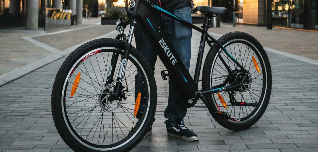 Ein Mann steht hinter einem dunklen E-Bike, rechts und links sind hohe Gebäude zu sehen