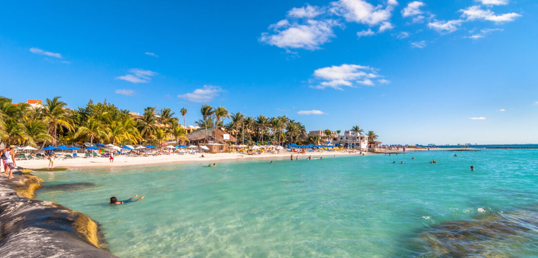 Badende im flachen Wasser an einem tropischen, touristischen Sandstrand mit Palmen und Badeliegen