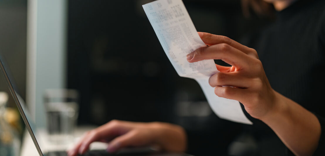 Eine Frau sitzt am Laptop und hält eine Quittung in der Hand