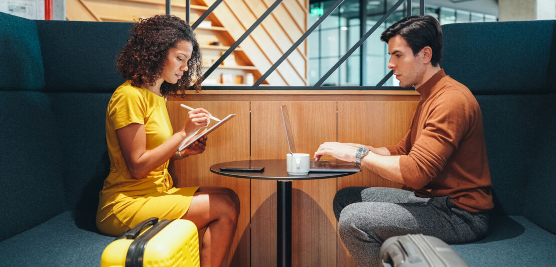 Ein Mann und eine Frau mit Koffern arbeiten an mobilen Geräten an einem Tisch in einer Flughafenlounge