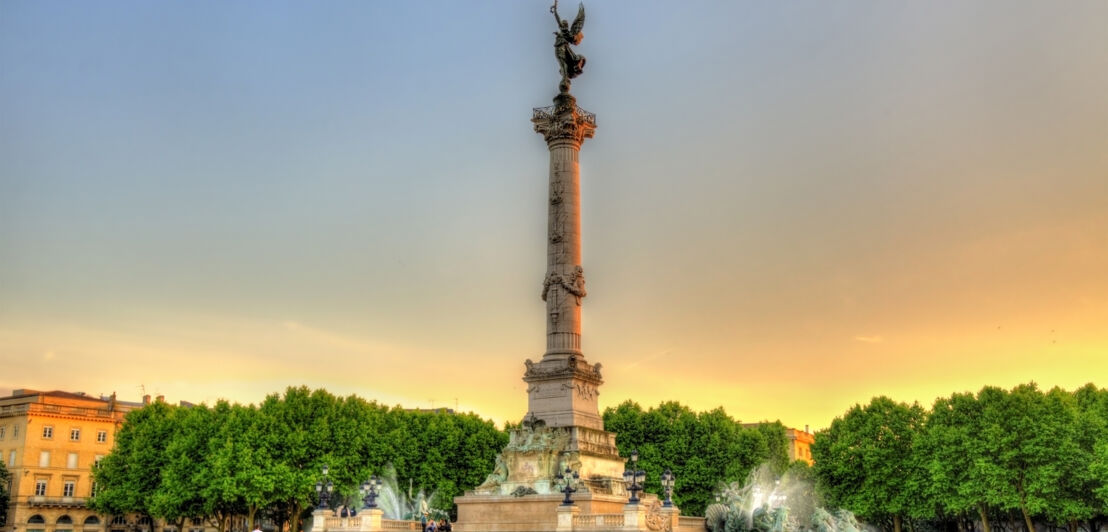 Eine Gedenksäule mit Engelsfigur auf einem großen Platz mit grünen Bäumen in der Abendsonne