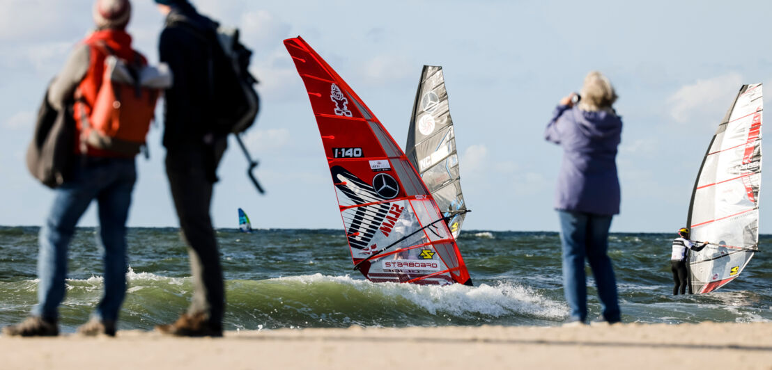 Personen am Strand beobachten Windsurfer bei einer Regatta im Meer