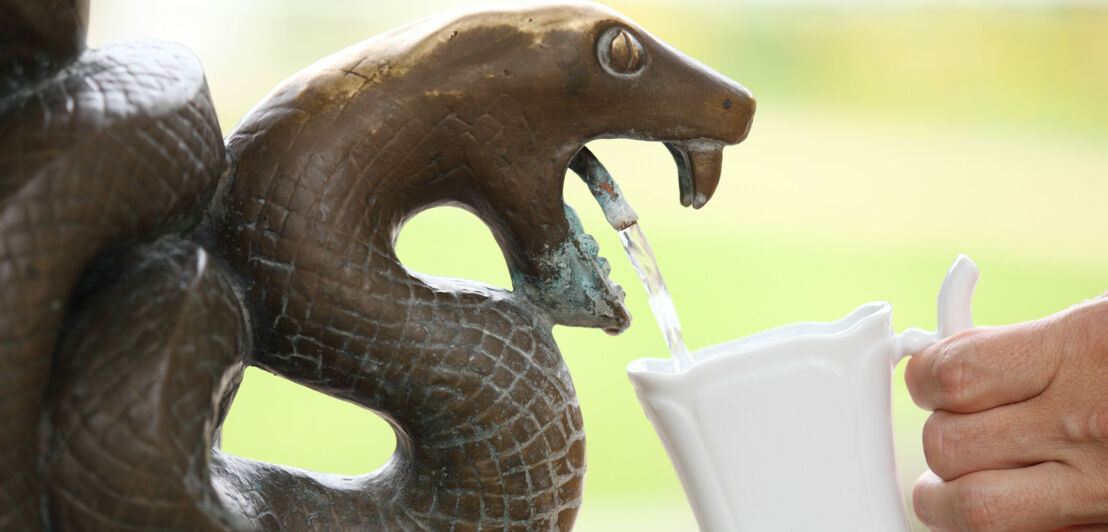 Eine Hand hält eine weiße Tasse unter einen Wasserhahn in Form einer Schlange