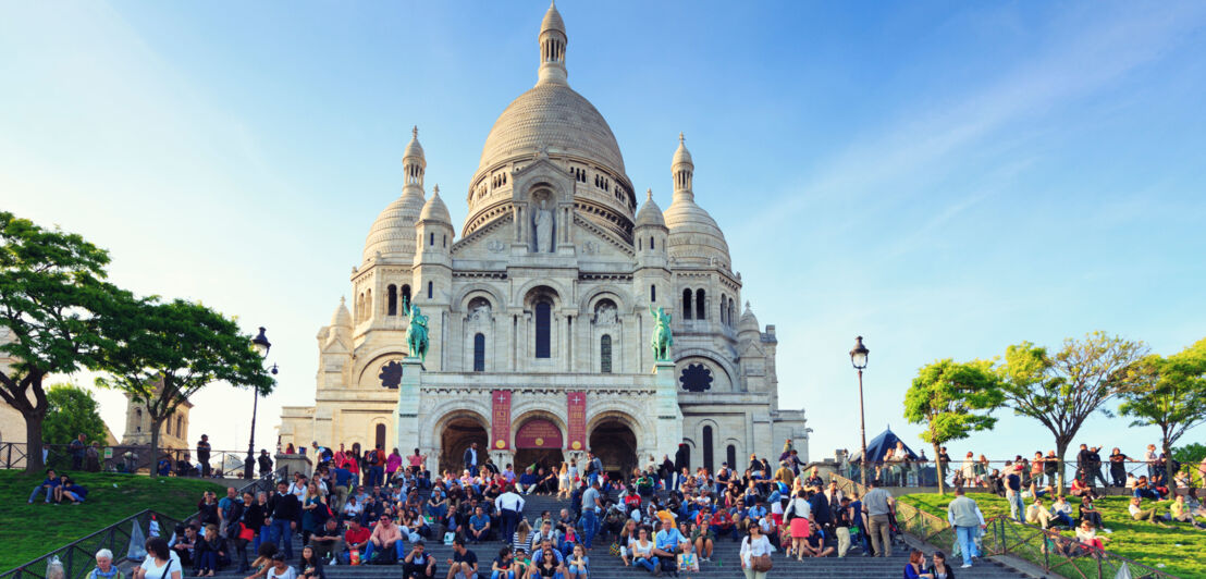 Sitzende Personen auf einer Treppe vor der Wallfahrtskirche Sacré-Coeur auf einem Hügel.