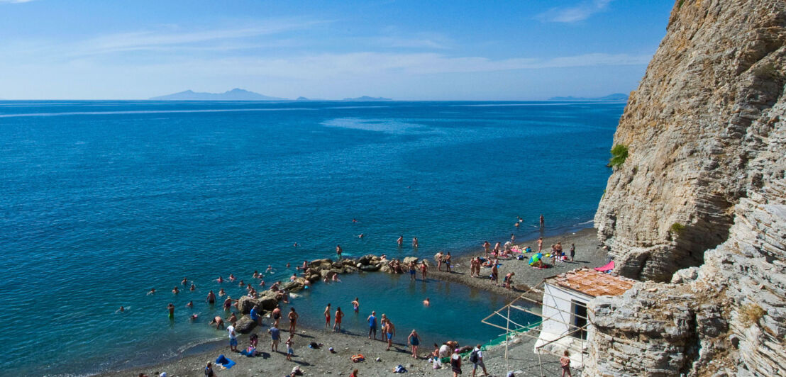 Personen baden in einem natürlichen Wasserbecken an einem Kiesstrand