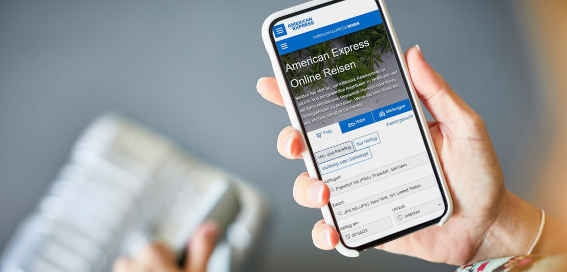 Ein Smartphone mit dem American Express Online-Reiseportal auf dem Display wird von einer Hand über einen Koffer gehalten.