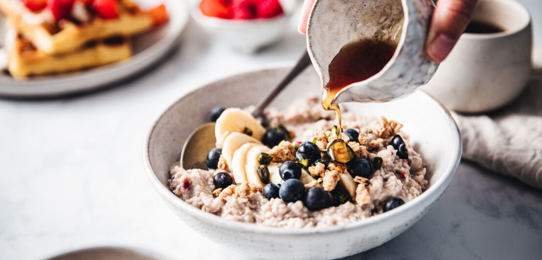 Eine Hand kippt Ahornsirup über eine Schüssel mit Porridge und Früchten.