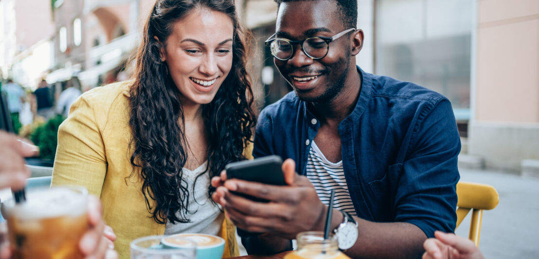 Ein junges Paar an einem Tisch in einem Straßencafé schaut gemeinsam auf ein Smartphone