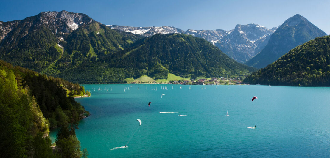 Ein großer See zwischen Bergen, auf dem Wasser zahlreiche Personen, die kitesurfen