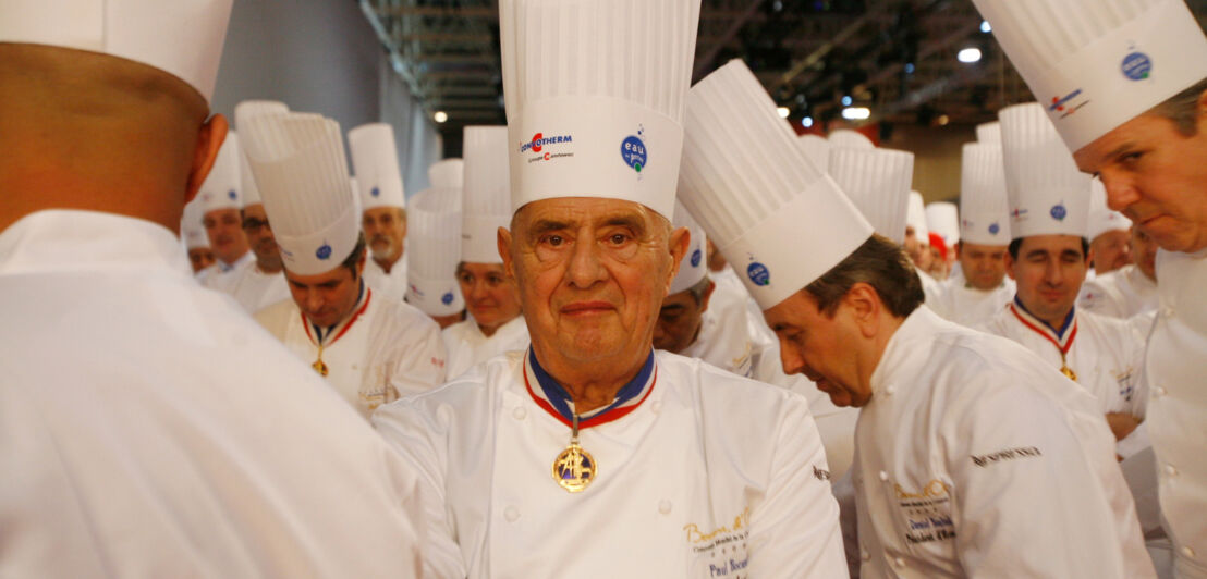 Porträt von Paul Bocuse in Kochuniform inmitten weiterer Köche in einer Halle