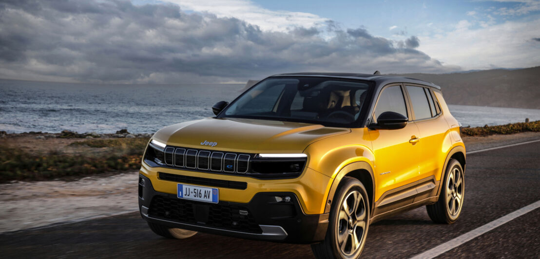 Ein goldgelbener Jeep Avenger fährt am Meer eine Straße entlang.