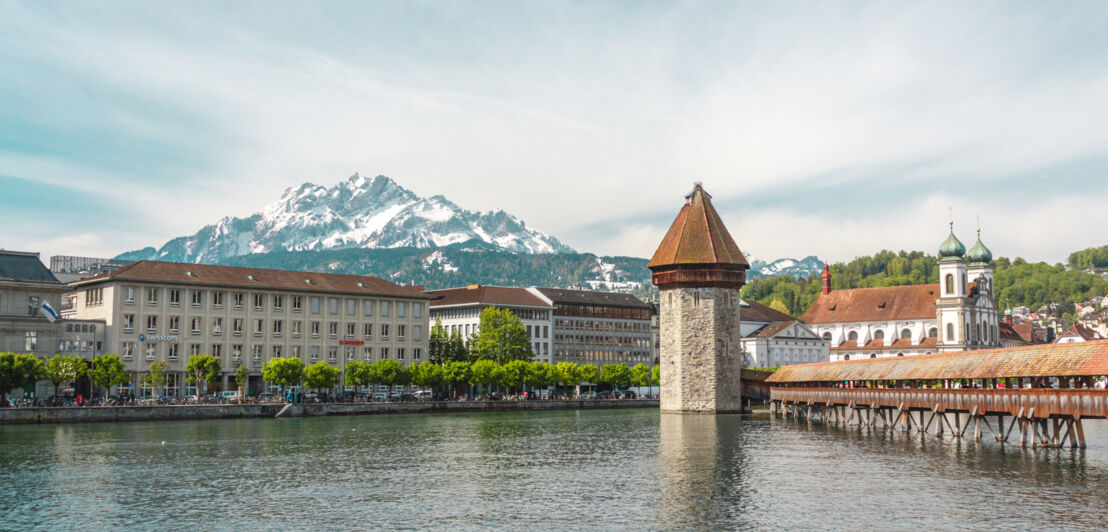 Eine Holzbrücke, ein Wasserturm, eine Stadtansicht und Berge im Hintergrund.