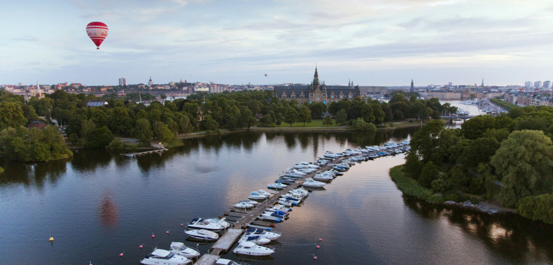 Blick über eine Stadt mit einem Fluss, Booten an einem langgezogenen Anleger und einem Heißluftballon über den Dächern.