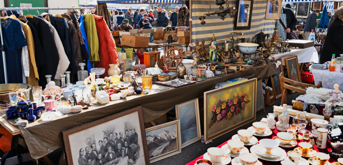 Stände mit Antiquitäten auf einem Trödelmarkt, im Hintergrund Personen.