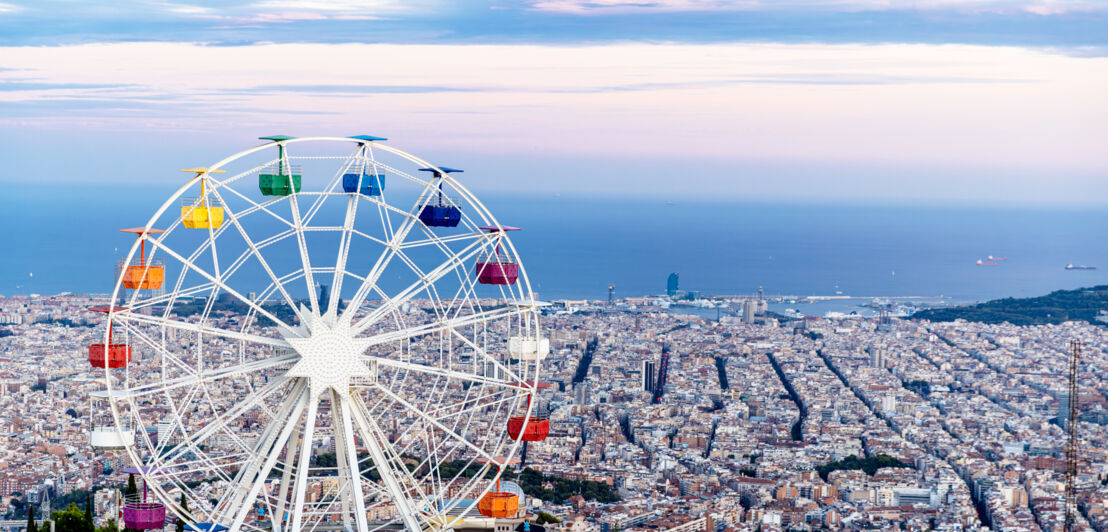 Ein Riesenrad auf einem Berg vor dem Stadtpanorama von Barcelona am Meer.