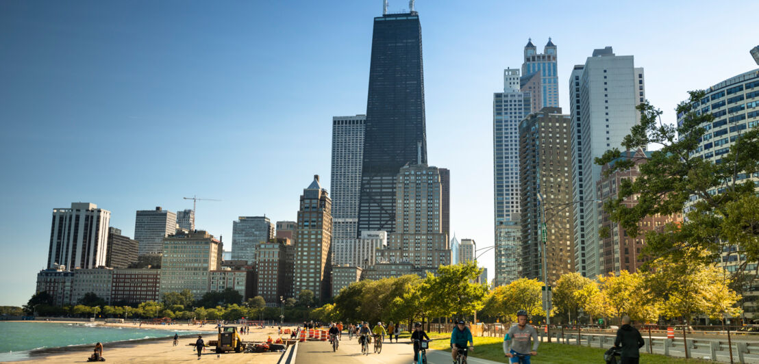 Radfahrende auf einem Weg am Seeufer vor der Skyline Chicagos.