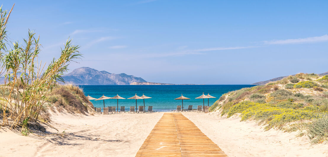 Weg durch Dünen zu einem hellen Sandstrand mit Sonnenliegen am türkisblauen Meer.