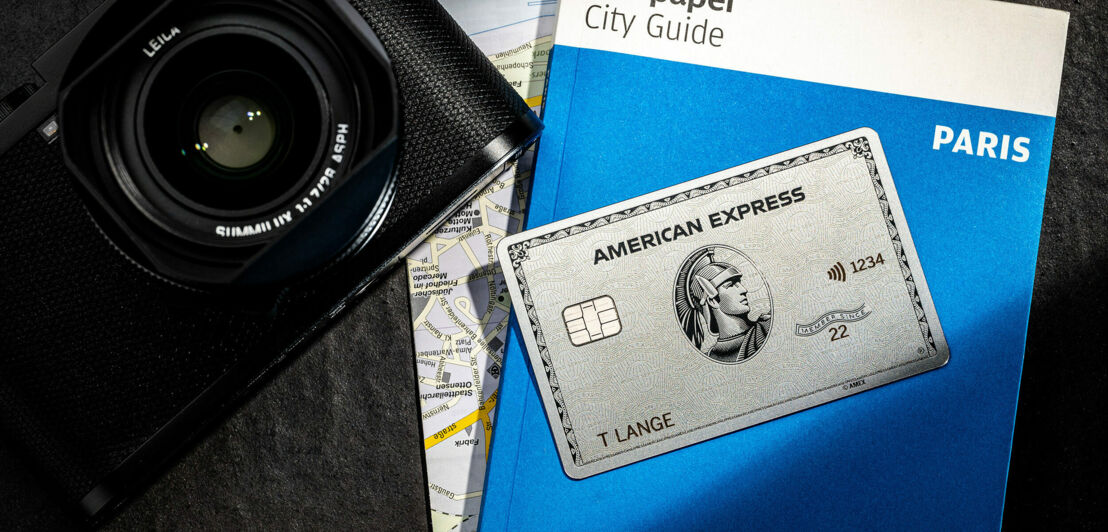 Eine American Express Platinum Card liegt auf einem blauen Reiseführer neben einer edlen, schwarzen Kamera