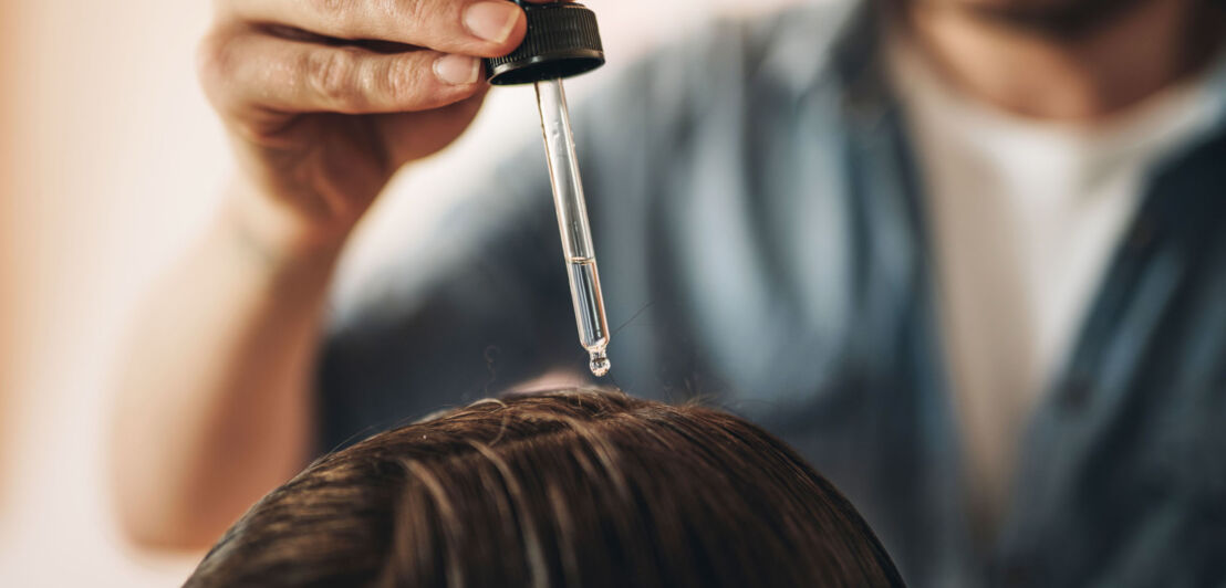 Nahaufnahme einer Hand mit Pipette, die eine Flüssigkeit auf den Haaransatz einer anderen Person träufelt.