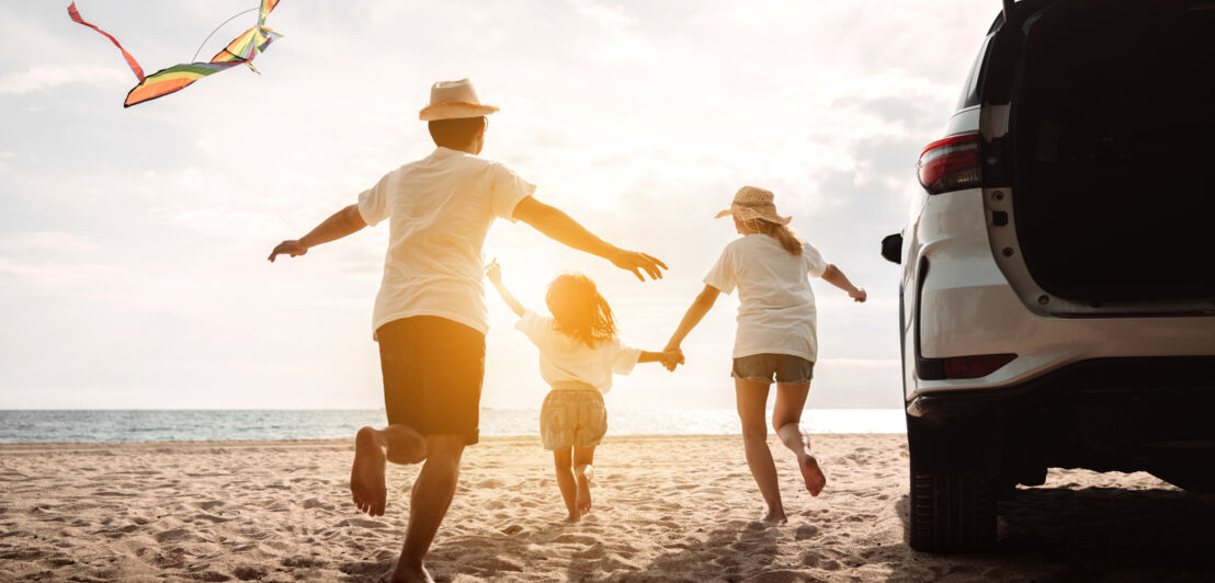 Rückansicht eines Mannes, eines Kindes und einer Frau in Shorts und weißen T-Shirts, die an einem Strand in Richtung Meer laufen, rechts neben ihnen ein weißes Auto von hinten im Anschnitt, am Himmel ein fliegender Drache.