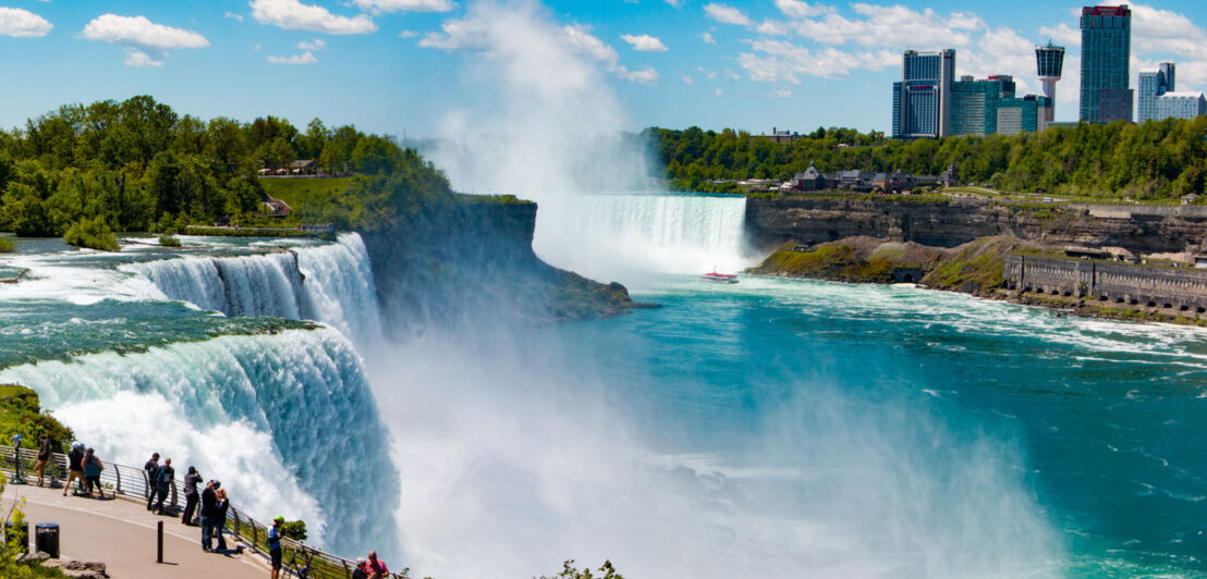 Panoramaaufnahme der Niagarafälle mit Personen am Ufern, im Hintergrund einige Hochhäuser.