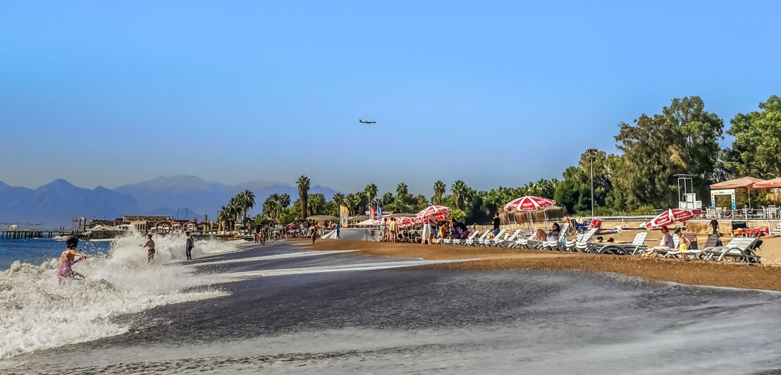 Kleiner Sandstrand mit Sonnenliegen und einigen Personen am Wasser, im Hintergrund Bäume und eine Gebirgskette.