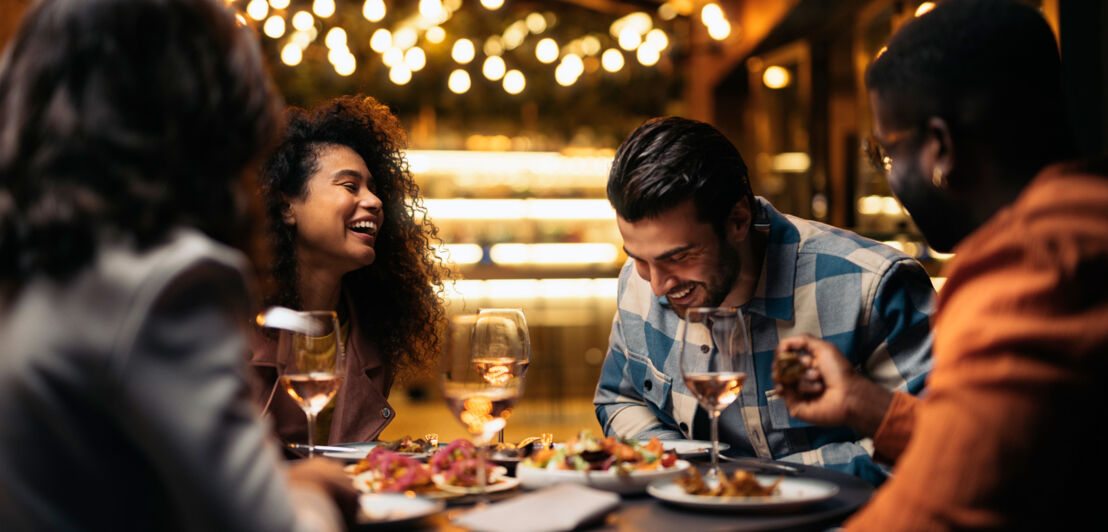 Vier junge Personen sitzen gut gelaunt beim Abendessen an einem runden Tisch in einem gehobenen Restaurant.