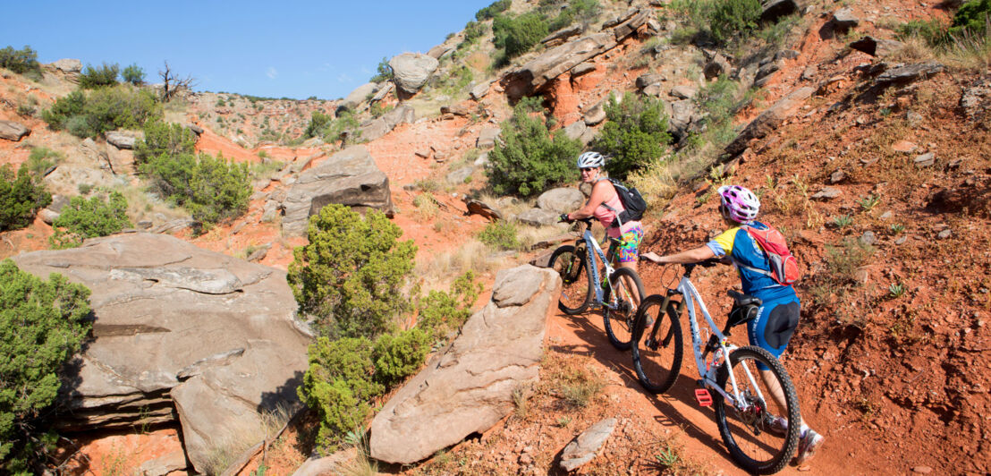 Zwei Frauen mit Mountainbikes in einer roten Wüstenlandschaft mit Felsbrocken und grünen Sträuchern.