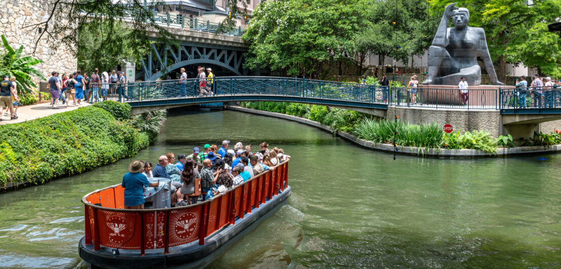 Ausflugsboot auf einer begrünten Wasserstraße vor einer Brücke mit Steinskulptur am im Stadtzentrum San Antonios.