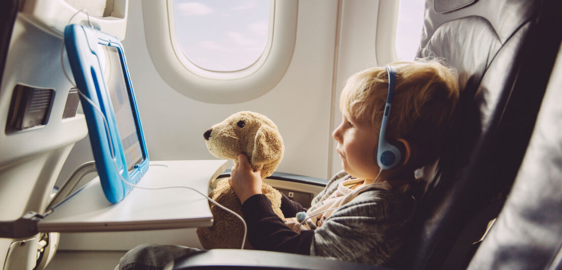 Ein kleiner Junge mit Kopfhörern in einem Flugzeugsitz schaut mit seinem Plüschhund einen Film auf einem Tablet.