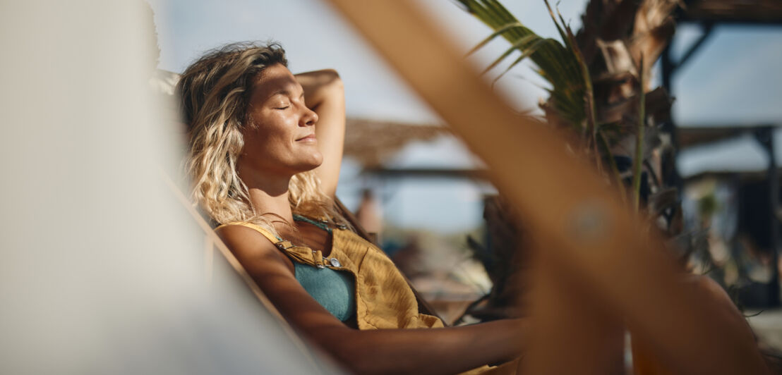 Nahaufnahme einer lächelnden Frau, die mit geschlossenen Augen die Sonne in einem Liegestuhl am Strand genießt