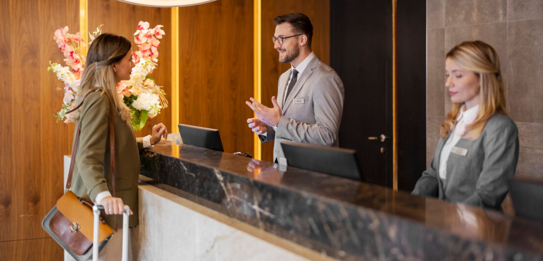 Eine Frau mit Koffer im Gespräch mit einem Mitarbeiter an einer Hotelrezeption.