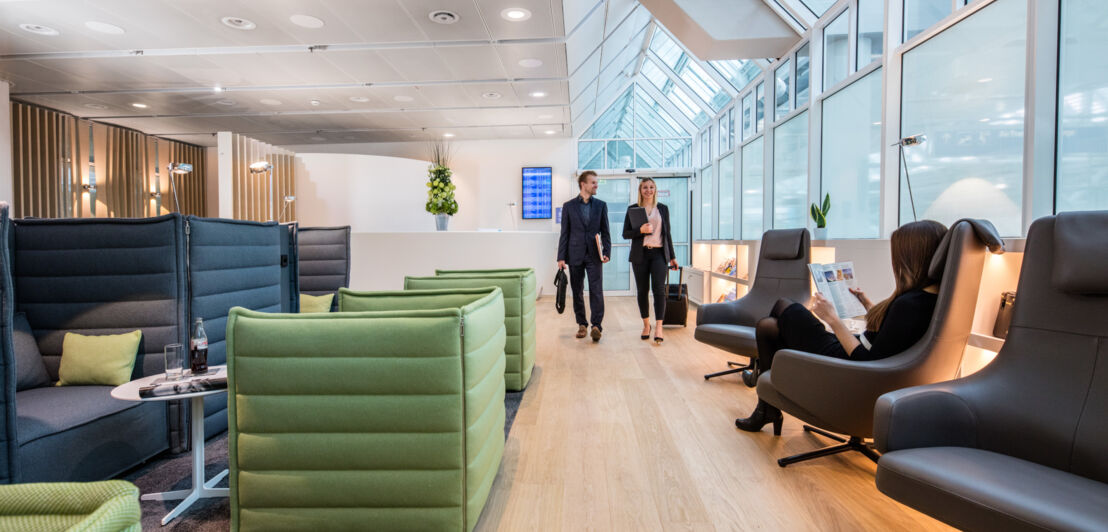 Mehrere Personen in einer modernen Flughafen-Lounge mit Sesseln vor einem Panoramafenster.